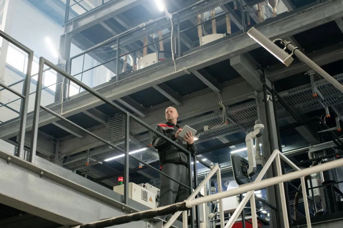 A plant leader with a tablet standing on a mezzanine, looking out over a multi-level industrial facility