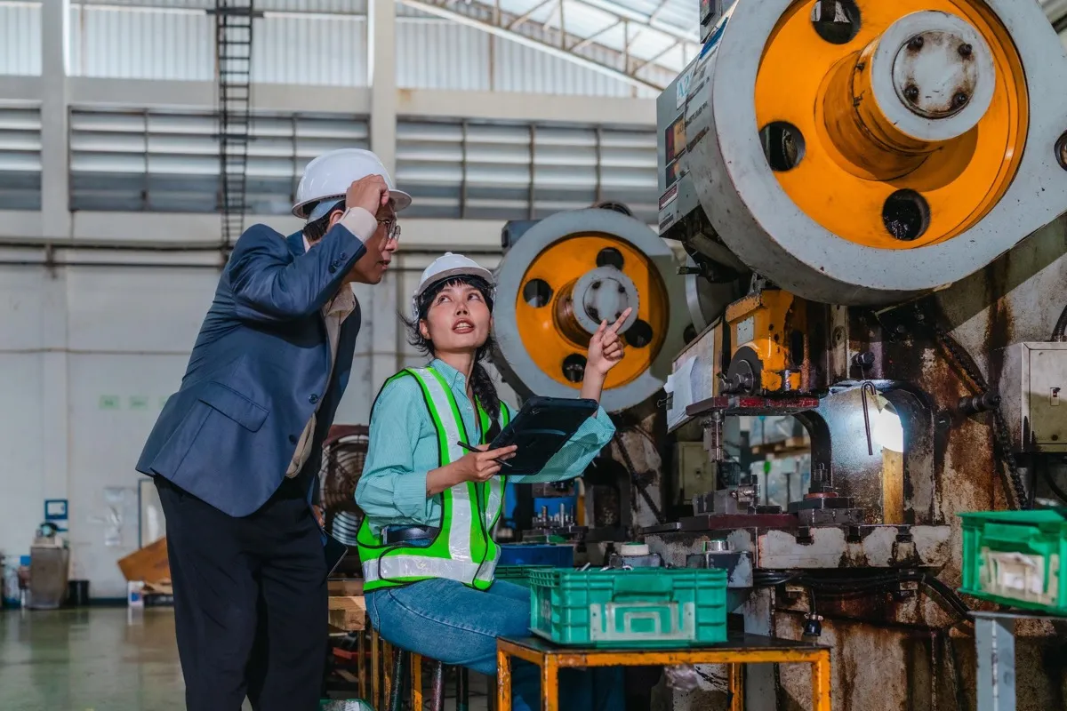 A maintenance and reliability leader and a technician in hard hats inspecting a press machine on the plant floor with a tablet