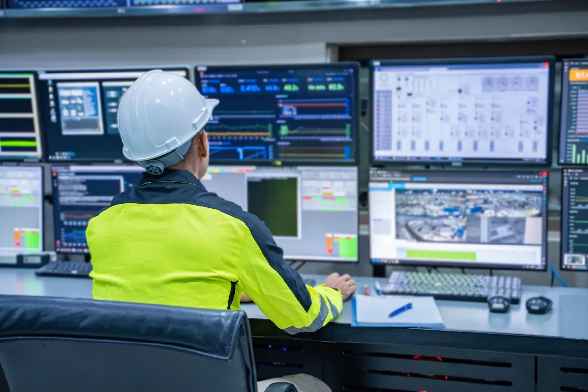 A plant operator in a hard hat and hi-vis jacket watching a control room wall of plant monitoring screens