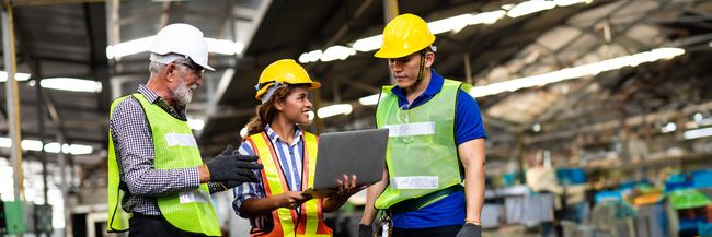 Three workers wearing hard hats on a manufacturing shop floor, holding a laptop